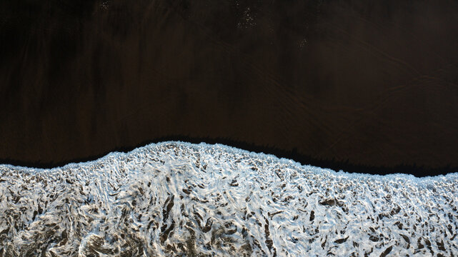 Aerial view of dark black sand and white sea foam from crashing waves along the shoreline in Piha, Auckland, New Zealand.