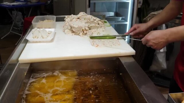 close-up shot of a street food vendor expertly shaping fresh fish paste on a white cutting board and sliding it into a vat of bubbling hot oil. The video captures the traditional process of making han