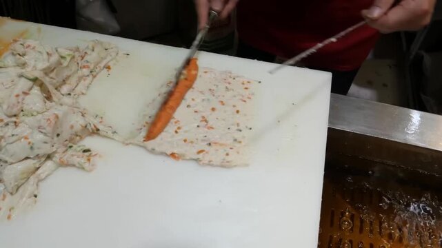 High-angle close-up of a street food vendor skillfully spreading a layer of bright orange fish roe or spicy seasoning over fresh fish cake paste on a white cutting board.