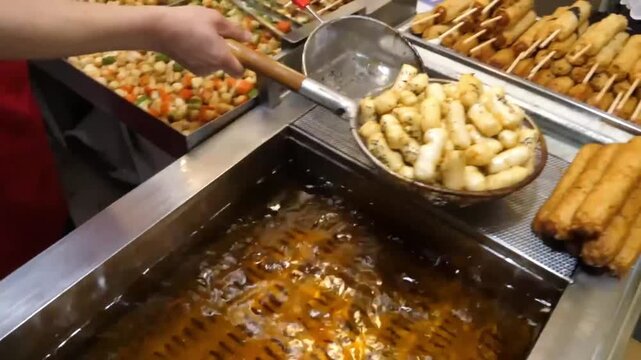 close-up shot of a street food vendor using a metal strainer to lift freshly deep-fried, chewy Korean rice cakes (tteokbokki) out of a hot oil boiler. The cylindrical rice cakes are crispy and coated 