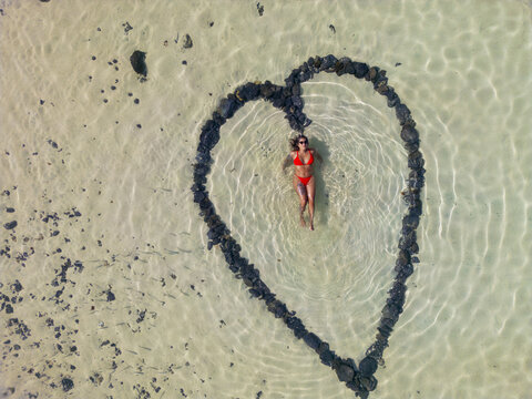 Aerial view of a woman in a red bikini floating inside a heart-shaped stone formation in the shallow clear water of El Cotillo, Canary Islands, Spain.