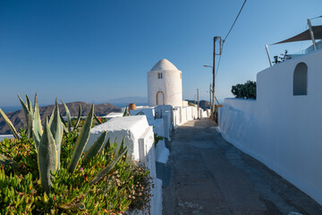 Quiet paved walkway with white Cycladic buildings, small chapel dome, and agave plants overlooking the Aegean Sea on Santorini, Greece. © Andrei Tomas