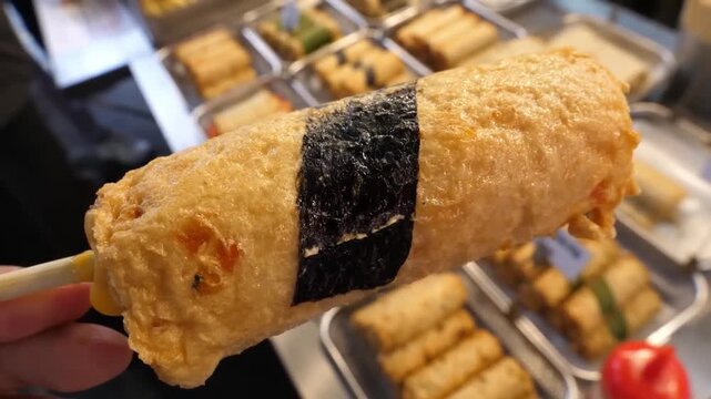 A close-up shot of a traditional Korean handmade fish cake (Eomuk or Odeng) on a wooden skewer. This popular street food snack is shown with a savory melted cheese center and a seaweed wrap, held over