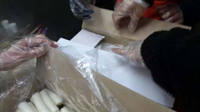 A top-down view of a cardboard shipping box lined with plastic, filled with rows of fresh, white, cylindrical Korean bar rice cakes (Garaetteok). These chewy rice cakes are a staple ingredient for pop