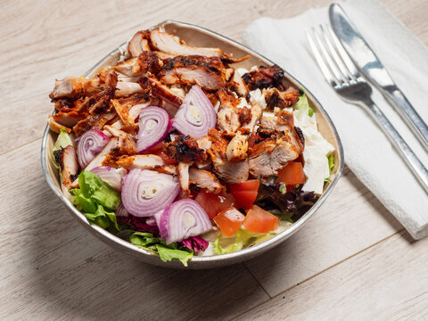 Grilled meat, onions, tomatoes, and lettuce in a bowl. Silverware rests on a napkin beside. Light wooden surface background. Tasty chicken salad meal serving,