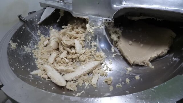 A high-angle close-up shot of a professional stainless steel bowl cutter machine processing thick slabs of raw fish paste (surimi) in a food manufacturing facility. This stage of Korean fish cake (Eom