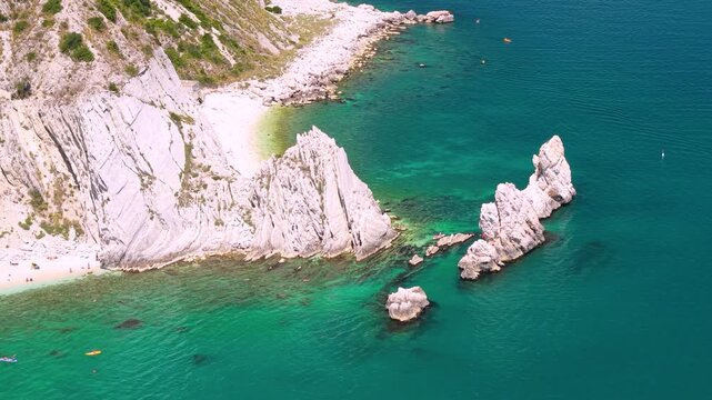 Sirolo Italy 4k drone 70mm telephoto of Two Sisters beach Due Sorelle faraglioni. High angle overflight of rocky limestone stacks with canoes in tropical turquoise sea water luxury travel b-roll