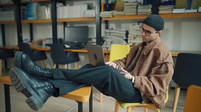 Student studies in a library using a laptop with books stacked on shelves nearby during the day