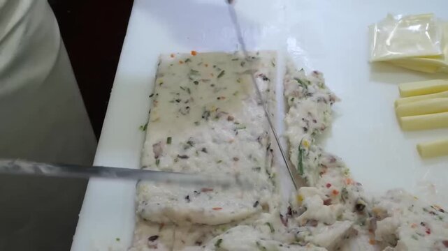 chef skillfully cuts and shapes fresh Korean fish cake paste (Eomuk) mixed with vegetables on a white preparation surface. This authentic street food preparation shows the traditional technique of cra