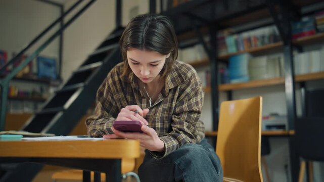 Young person studies in a library while using a phone and sitting at a table on a weekday afternoon