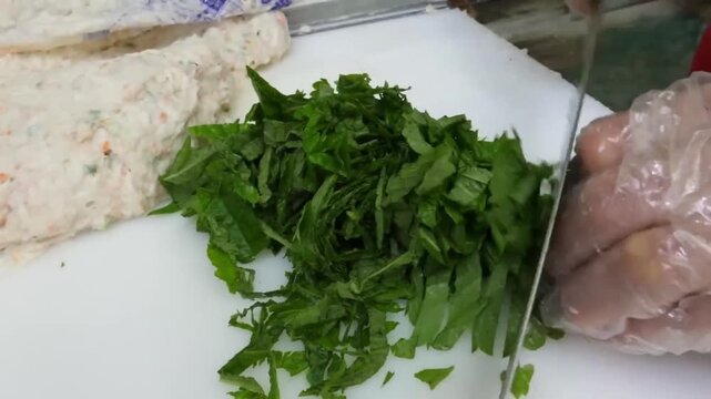 close-up shot of a chef using a knife to chop fresh green perilla leaves on a white cutting board next to fish paste mixture.