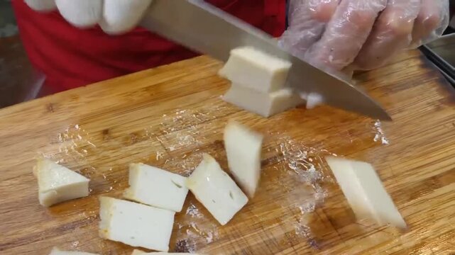 Close-up of a chef wearing gloves slicing a long piece of traditional Korean fish cake (eomuk) lengthwise on a wooden cutting board in a kitchen.