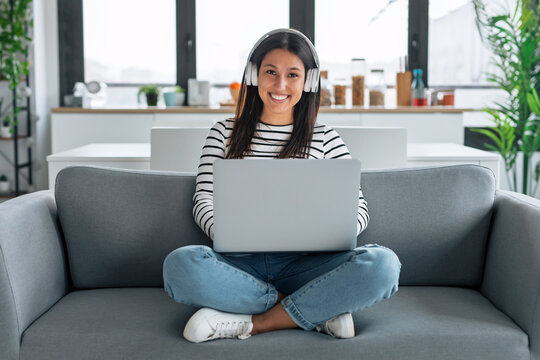 Happy woman listening to music with headphones while using laptop sitting on the couch at home