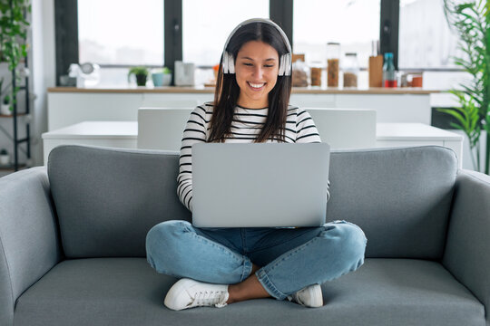 Happy woman listening to music with headphones while using laptop sitting on the couch at home