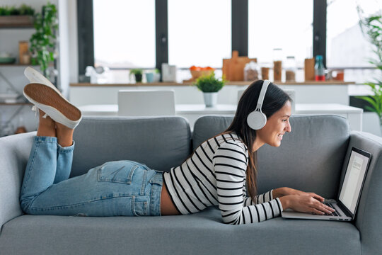 Smart woman listening to music with headphones while using laptop lying on the couch at home