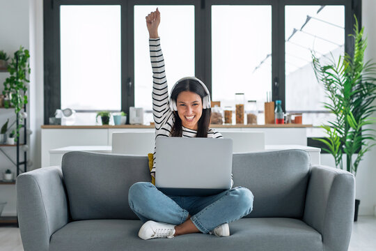 Smiling woman with headphones celebrating while using laptop sitting on the couch at home