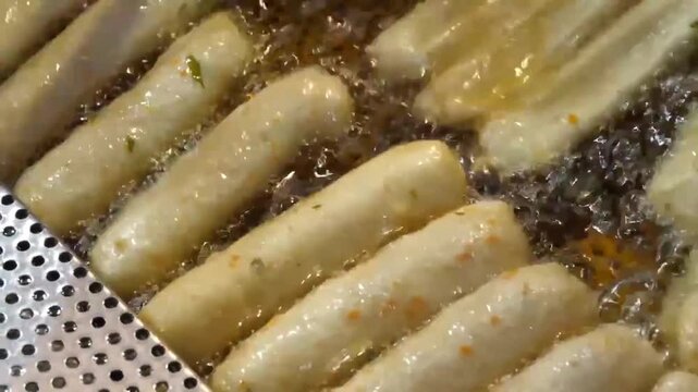 Close-up of traditional Korean fish cakes (eomuk or hot bar) being deep-fried in hot oil until golden brown. A chef uses tongs to flip the long, cylindrical fish cakes, showcasing a popular Korean str