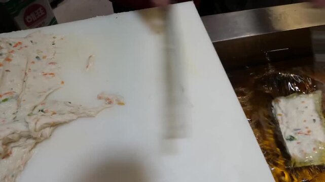 High-angle close-up footage of a skilled street food vendor meticulously shaping fresh fish cake paste (surimi) mixed with vegetables on a white preparation board. The scene captures the traditional a