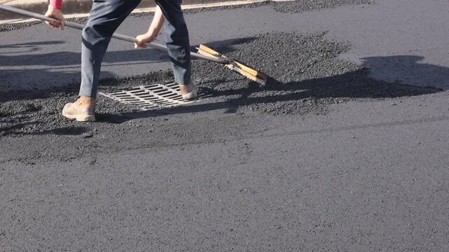 Worker uses rake to spread asphalt over street near drain while others assist in repair work