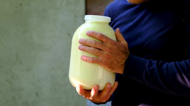 A man stands outdoors near a village house holding a large three liter jar of fresh homemade milk. Traditional countryside lifestyle, farm products and local dairy food.