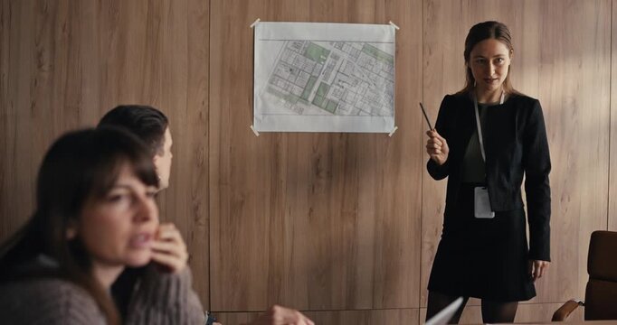 A woman presents project plans to a group in a meeting room while showing maps on the wall at midday