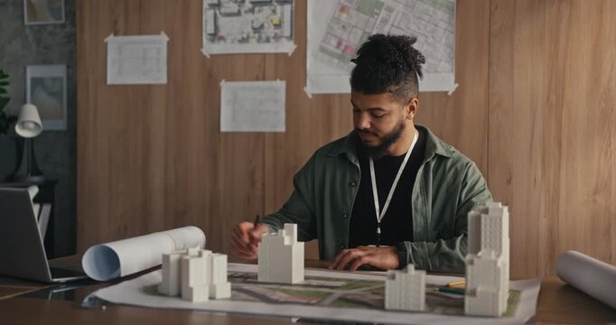 A designer creates a city model on a table in an office. Plans and maps are on the wall in the background