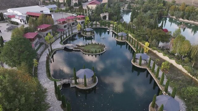 An aerial drone view showing a beautiful resort park with a calm lake, elegant gazebos, stone pathways, and lush cypress trees reflecting in the water during sunset.


