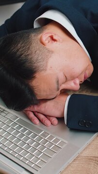 Bored sleepy Asian businessman worker working on laptop computer, leaning on hand falling asleep at office desk. Exhausted tired freelancer workaholic middle-aged man. Employment, occupation, workless
