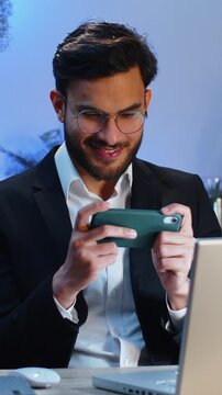 Indian businessman playing on smartphone mobile application celebrate winning racing or shooter game at office desk workplace. Professional manager freelancer man enjoy testing good work of new app