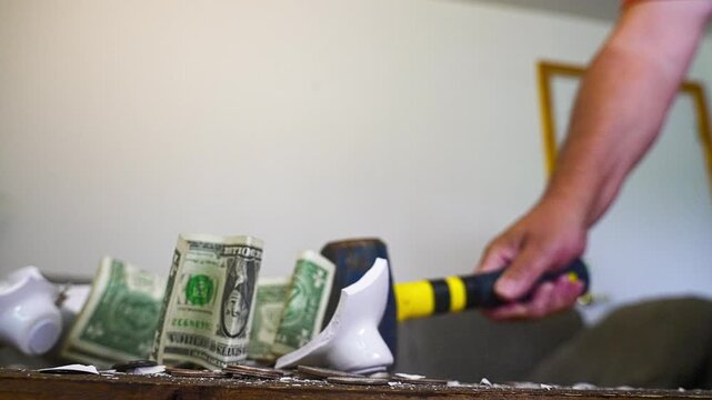 Slow motion footage showing a hand smashing a white ceramic piggy bank with a hammer. Coins and dollar bills explode from the broken pieces