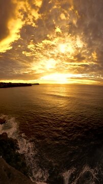 Slow-motion wide-angle video of a sunrise over the ocean along the Sydney coastline, with a coastal cliff wall illuminated by soft morning light and calm sea conditions, Australia.