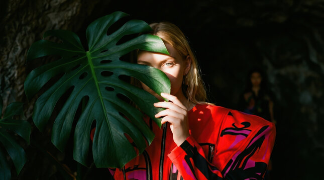 Fashion model poses with large leaf in vibrant outfit during outdoor photoshoot in natural setting