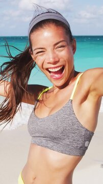 Happy sporty woman taking selfie with phone laughing after running on beach. Girl taking fitness selfie after workout on summer vacation living healthy lifestyle having fun. VERTICAL SLOW MOTION