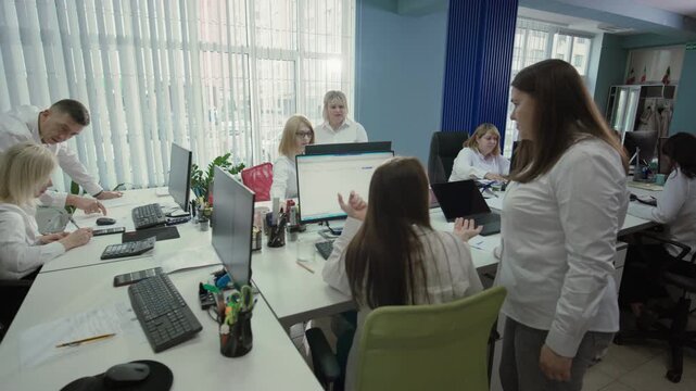 Wide shot of Caucasian employees in an open office with natural light. Several women work at computers, assisted by a man. 4K.