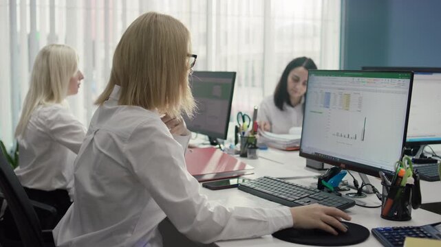 Commercial close-up shot from behind of Caucasian female employee working at computer. A graph is displayed on the screen while female colleagues work in the background. 4K.