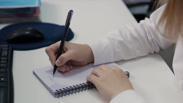 Extreme close-up on a desk, showing the hands of an employee writing notes in a spiral notebook. The keyboard and a mouse pad are visible. 4K.