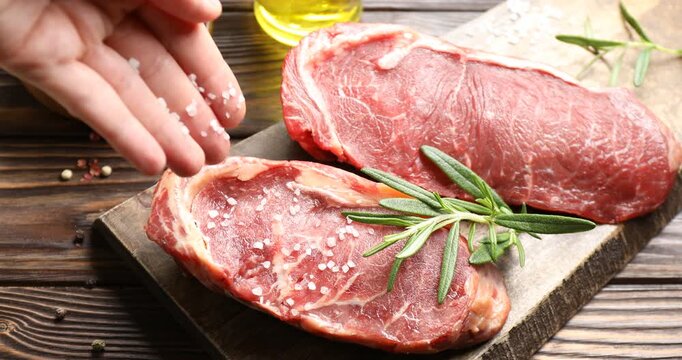 Woman adding salt to raw beef steak at wooden table, closeup
