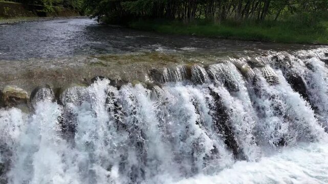 Water rushes over rocks in a natural cascade. White foam swirls as the stream tumbles. Green trees frame the rushing river. Calm forest surroundings echo the water's motion