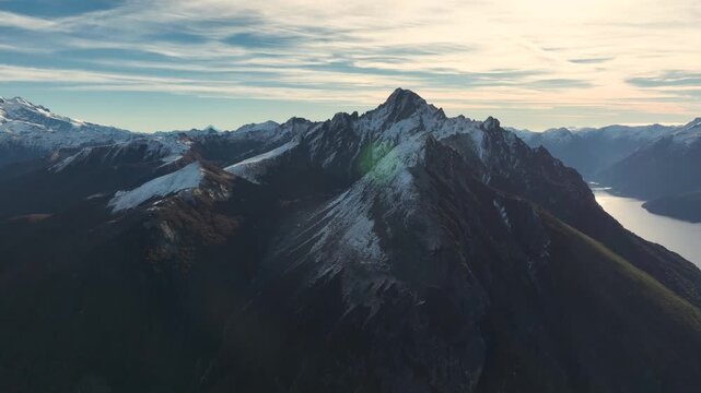 Aerial drone view of a jagged, snow-dusted mountain summit next to a glowing water body under a dramatic evening sky.