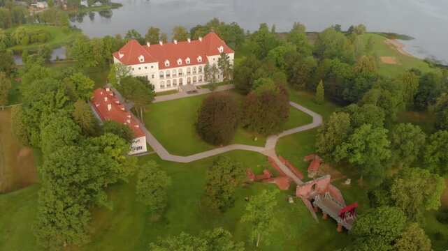 An aerial shot showcasing the beautiful 17th century Biržai Castle (Lithuanian Biržų pilis) complex in northern Lithuania. The white palace with its distinctive red roof is situated amidst lush green 