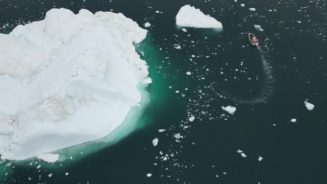 Aerial view of a small boat navigating through dark waters near a massive iceberg in Ilulissat, Avannaata Municipalit, Greenland.