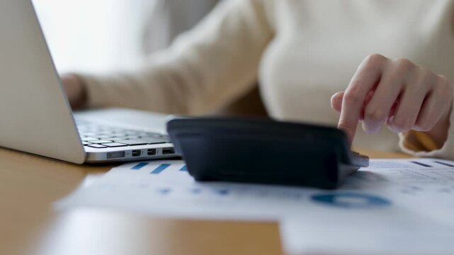 Person using a calculator and laptop, reviewing printed financial charts and reports on a desk, performing budgeting and data analysis in an office setting