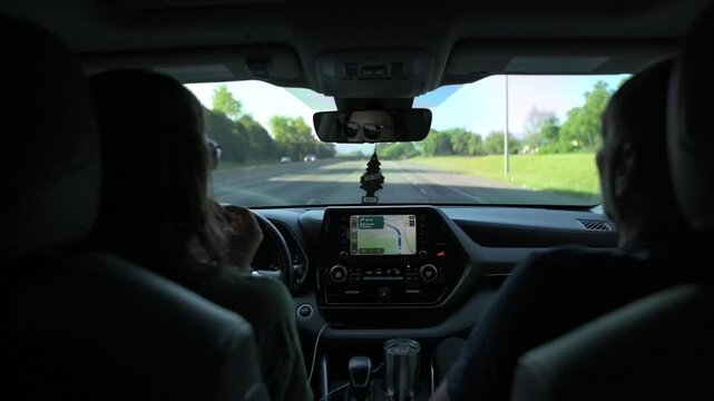 Two individuals seated in a car, with one person driving and the other navigating using a dashboard display, surrounded by lush greenery along the road on a bright sunny day
