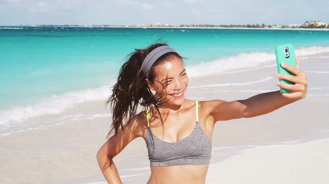 Beautiful young woman taking selfie on beach using smart phone. Video is unretouched and model without makeup. Raw footage.