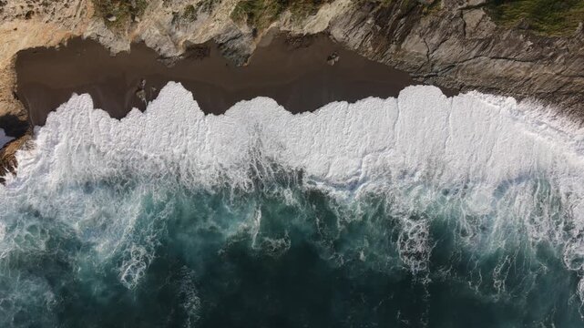 Aerial view of Playa de la Cueva showing turquoise ocean waves crashing against the dark sandy beach and rocky cliffs in Spain.