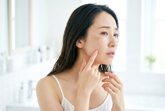 洗面所で鏡を見ながらシミや肝斑やそばかすを気にする30代日本人女性｜美容と肌悩みのイメージ