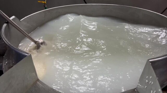 Worker stirring soaked rice in a stainless steel processing tank during food production. Rice preparation process in a food manufacturing facility before cooking and further processing.