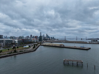 Obraz premium San Francisco skyline and Bay Bridge on a cloudy day, showing modern buildings, urban waterfront development, and industrial piers alongside Mission Bay