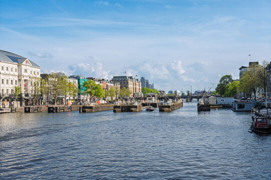 A beautiful sunny day view of the Amstel River in Amsterdam, Netherlands, featuring historic buildings, canal infrastructure, and calm water under a bright blue sky in spring.