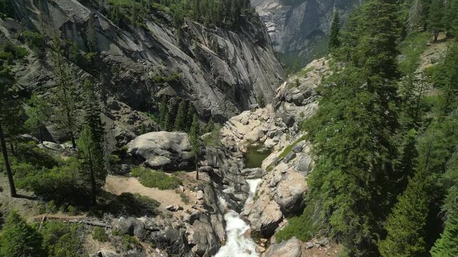 Aerial shot of a river flowing through Yosemite Valley with a cascading waterfall surrounded by granite cliffs and dense forest in California.